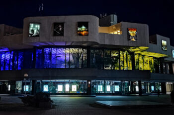 Zdjęcie. Oświetlony budynek Kieleckiego Centrum Kultury nocą. Na pierwszym piętrze foyer oświetlone niebieskim i żółtym światłem scenicznym.