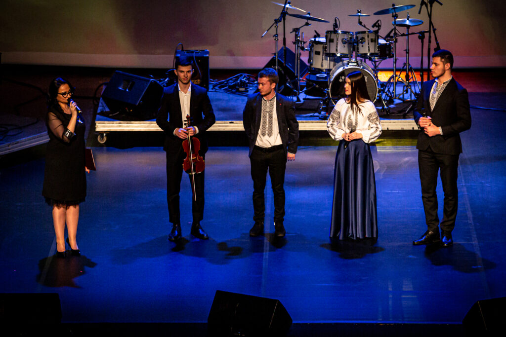 Zdjęcie. Na scenie stoi czwórka młodych muzyków: trzech mężczyzn i jedna kobieta w koszuli z niebieskim haftem. Po lewej prowadząca koncert Aneta Czaban