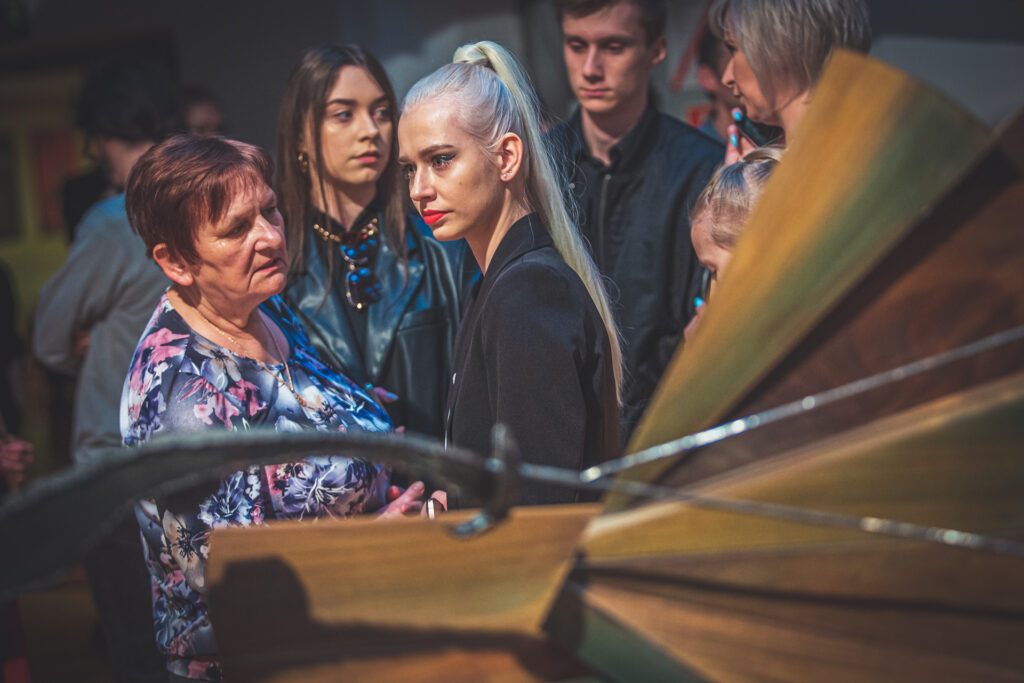 Zdjęcie. Publiczność wernisażu Na pierwszym planie dziewczyna z długimi prostymi włosami blond, spiętymi na czubku głowy w kucyk. Obok kobieta w średnim wieku w kwiecistej bluzce