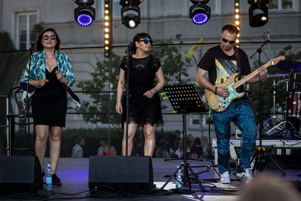 Zdjęcie. Trójka muzyków - dwie chórzystki i gitarzysta na scenie na rynku.