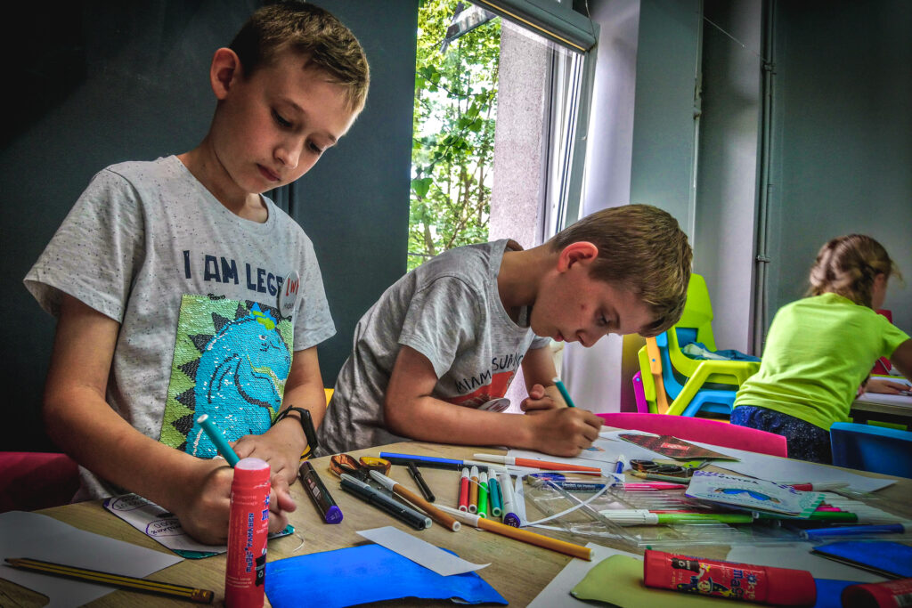 Zdjęcie. Dwaj chłopcy pracują przy stole w pracowni plastycznej. Obaj malują flamastrami.