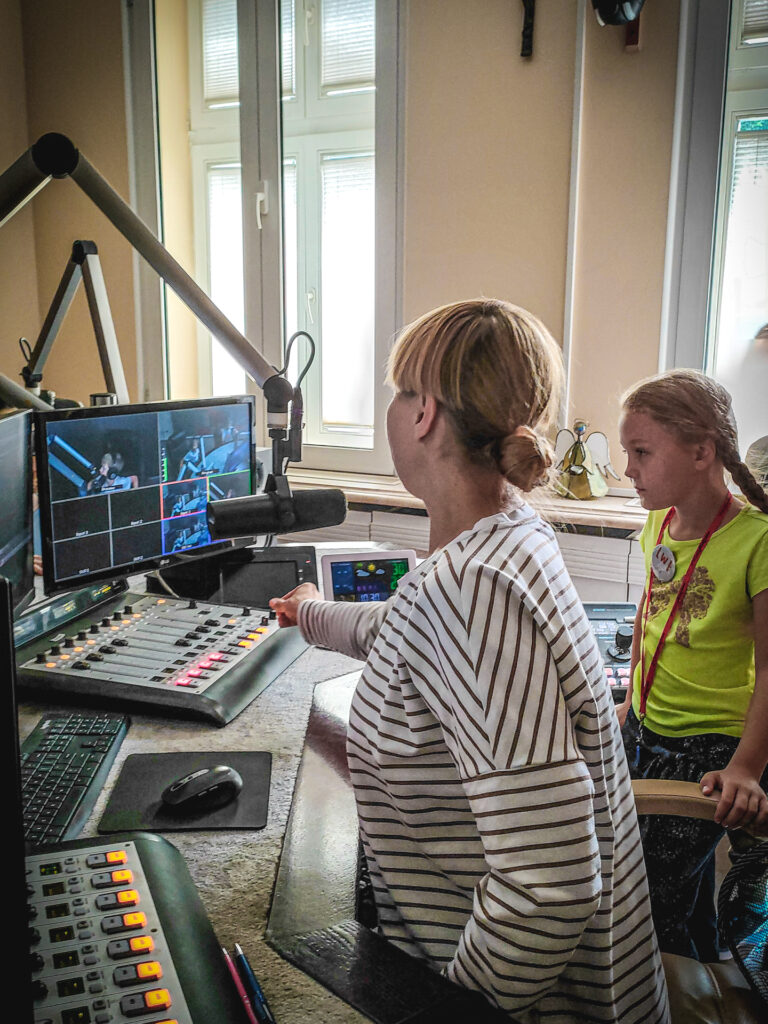 Zdjęcie. Dziewczynka przygląda się realizatorce audycji radiowej. Patrzy na komputery i miksery.