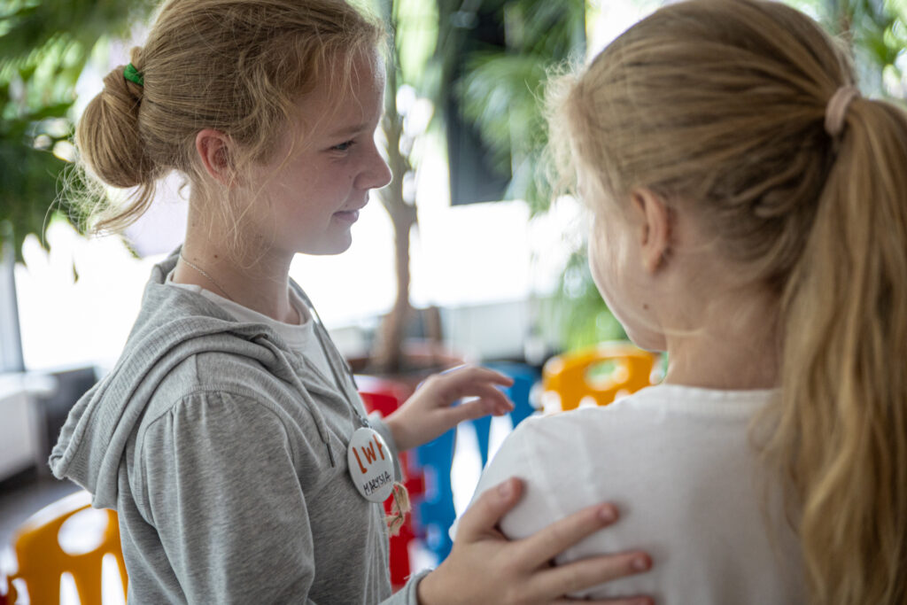 Zdjęcie. Dwie blondynki uśmiechają się do siebie. Jedna dotyka ręką pleców drugiej.