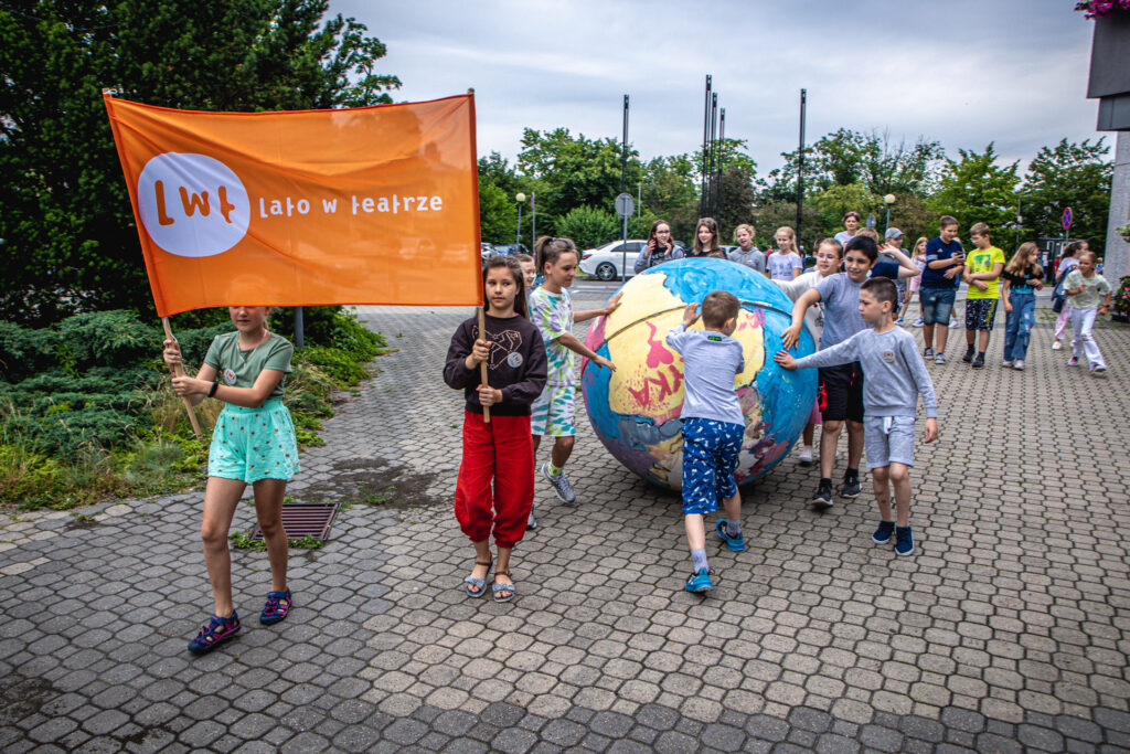 Zdjęcie. Grupa dzieciaków idzie w pochodzie. Dwie dziewczynki trzymają pomarańczowy transparent z napisem Lato w Teatrze. Za nim niewielka grupa pchająca dużą styropianową kulę z narysowanymi kontynentami. W tle pozostałe dzieci z warsztatów.