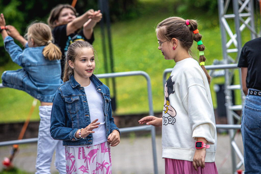 Zdjęcie. Dwie dziewczynki rozmawiają ze sobą. Jedna ma długie włosy spięte kilkoma kolorowymi frotkami.