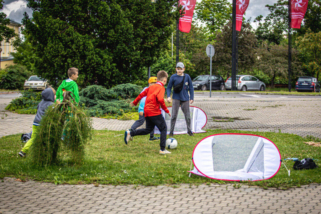 Zdjęcie. Plac przed KCK. Pięciu chłopaków gra w piłkę do małych bramek. Przy piłce chłopiec w czerwonym dresie.