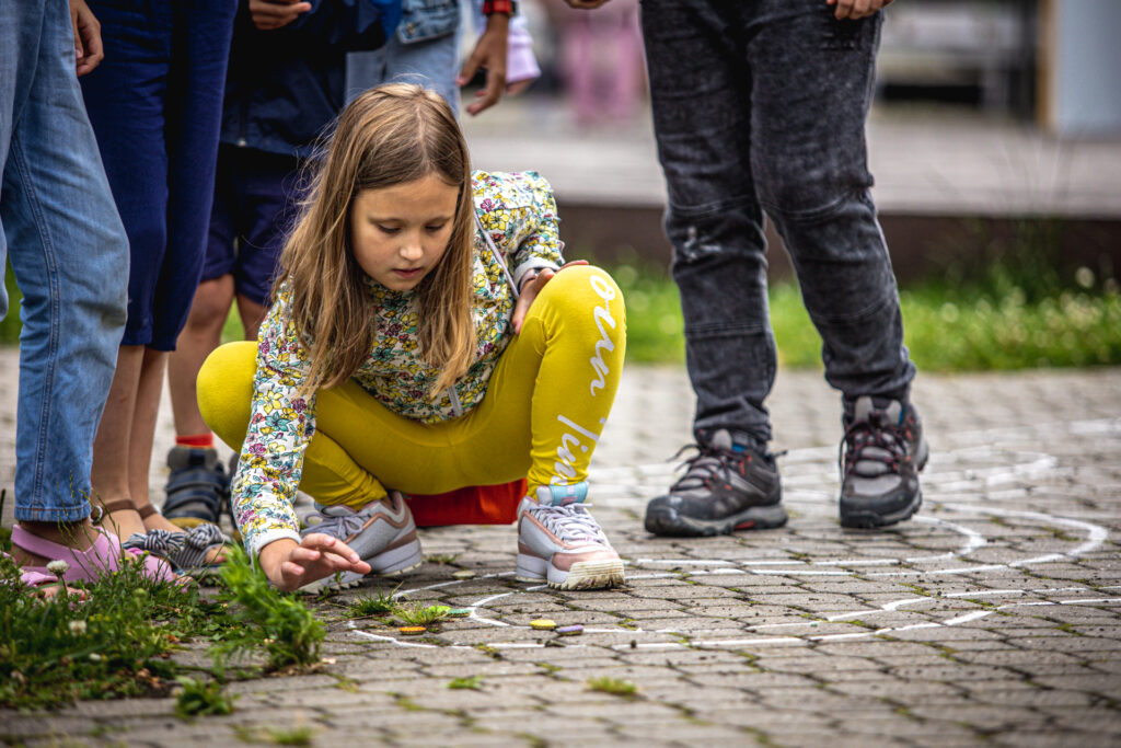 Zdjęcie. Blondynka kuca jednocześnie pstrykając kapslem po narysowanej kredą drodze.