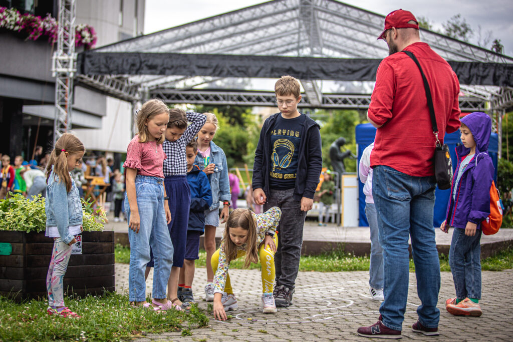 Zdjęcie. Dzieci grają w kapsel. Na kostce namalowana jest trasa. Dzieci pstrykają kapsle wypełnione kolorową plasteliną.