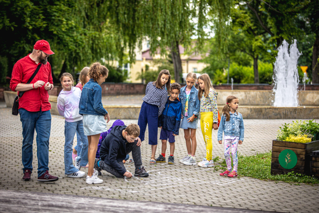 Zdjęcie. Plac przed KCK. Osiem dziewczynek przygląda się chłopakowi w okularach, który maluje kredą po kostce brukowej. Obok stoi wysoki postawny mężczyzna w czerwonej czapce bejsbolówce i czerwonej koszuli.