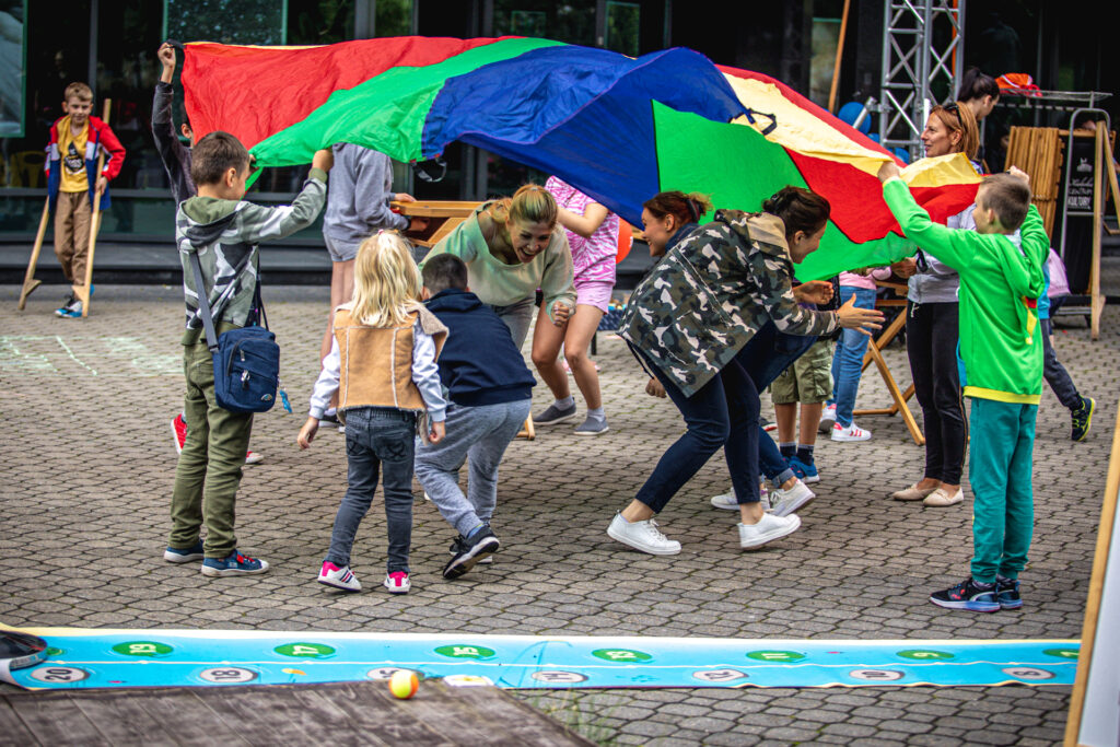 Zdjęcie. Grupa osób przebiega pod ogromną kolorową chustą, którą ruszają inne dzieci.