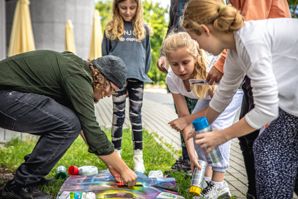 Zdjęcie. Dzieciaki pochylają się nad kolorową kartką papieru położoną na trawie. Jedna z dziewczynek trzyma w dłoni spray z niebieską farbą.