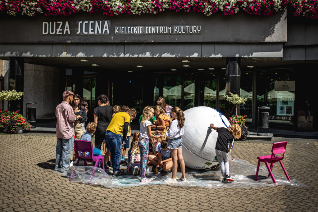 Zdjęcie. Teren przed Kieleckim Centrum Kultury. Dzieci malują duża styropianową kulę. Nad nimi napis na balkonie: Duża Scena Kieleckiego Centrum Kultury.