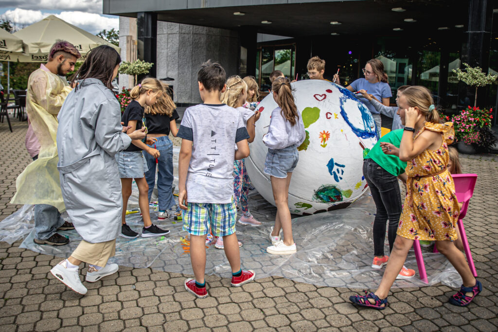 Zdjęcie. Plac przed KCK. Na dużej folii stoi ogromna styropianowa kula. Dzieciak stoją przy niej i malują ją na różne kolory. PObok dwoóka wolontariuszy Hayk z Armenii i Georgia z Grecji.