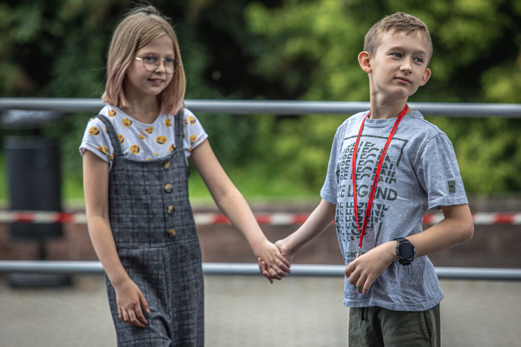 Zdjęcie. Para dzieciaków z warsztatów Lato w Teatrze. Dziewczynka o prostych włosach blond do ramion trzyma za rękę chłopca w szarej bluzce z czerwoną smyczą na szyi. Chłopiec odwraca głowę do tyłu.