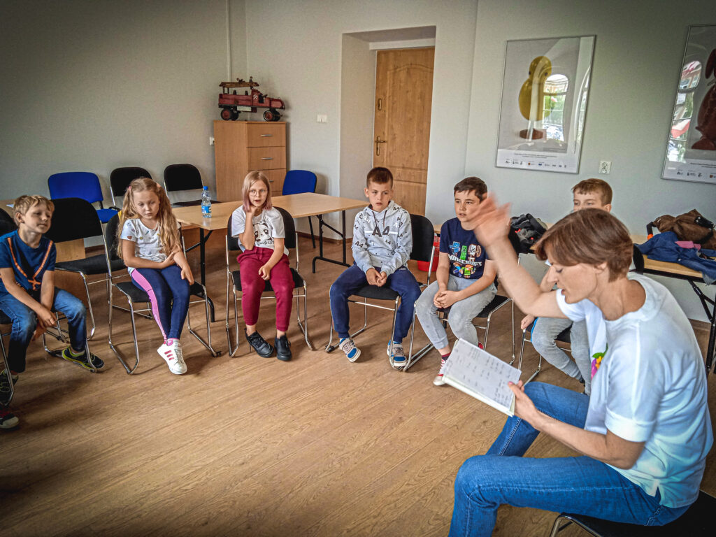 Zdjęcie. Grupa dzieci siedzi w kółku w sali konferencyjnej. Prowadząca Ewa Lubacz czyta coś w zeszycie.