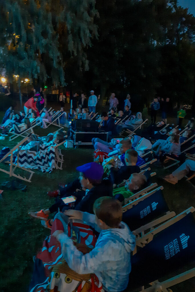 Zdjęcie. Późny wieczór. Trwa pokaz filmu podczas imprezy Kino na Osiedlach. Widzów oświetla niebieskie światło ekranu.