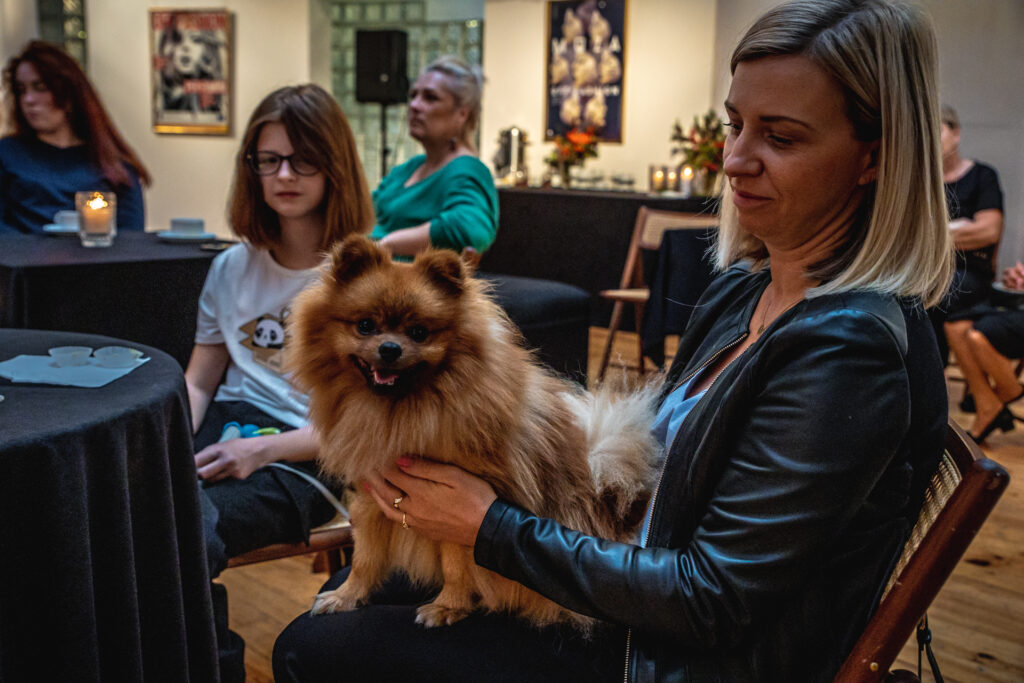 Zdjęcie. Blondynka o prostych włosach do ramion trzyma na kolanach niedużego psa o rudej sierści. To widzka spotkania z Dorotą Sumińską.