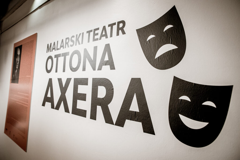 Zdjęcie. Biała ściana z napisem Malarski Teatr Ottona Axera oraz grafiką przedstawiającą smutną maskę teatralną.. W tle tablica informacyjna ze zdjęciem Axera.