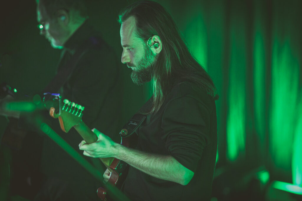 Zdjęcia. Giatrzysta z długimi włosami gra na instrumencie. Scenę oświetla zielone światło.