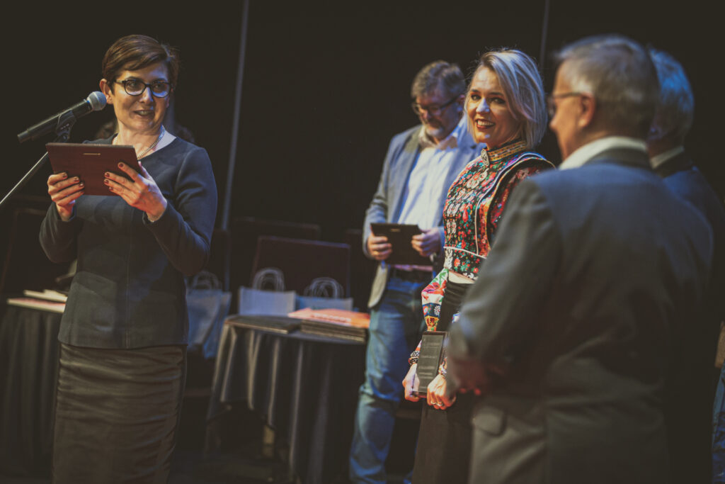 Zdjęcie. Kobieta w okularach i czarnej garsonce mówi do mikrofonu. To dyrektor KCK Augustyna Nowacka. Obok niej stoją uśmiechnięci panowie i panie. Finaliści świętokrzyskich eliminacji konkursu Mistrz Mowy Polskiej.