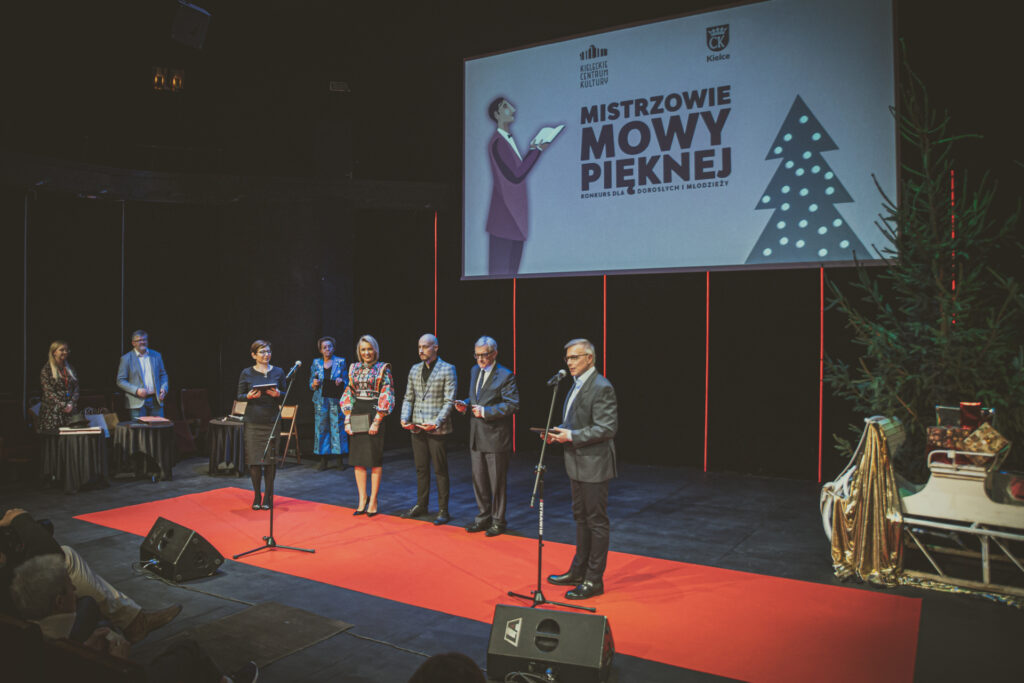 Zdjęcie. Mała Scena KCK. Na pierwszym planie czerwony dywan. Na nim stoi męćzyznza przed mikrofonem na statywie. W tle trójka ludzi: dwóch męćzyzn i jdna kobieta. Obok kolejna kobieta z książką w ręce.