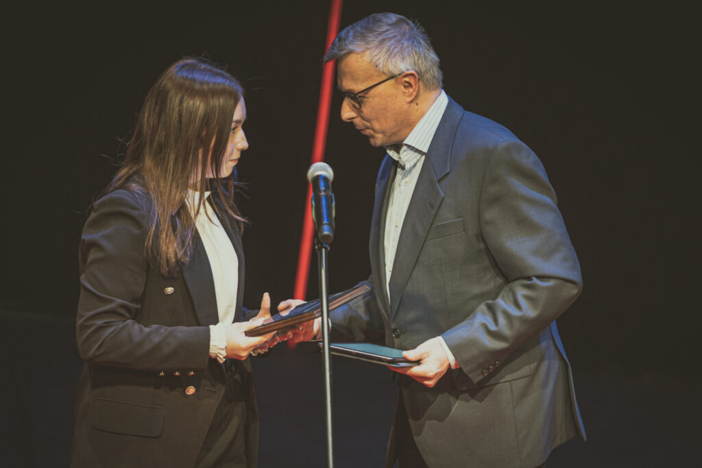 Zdjęcie. Męczyzna z siwymi włosami w okularach przekazuje nagrodę dziewczynie w jasnej bluzce i ciemnej marynarce.