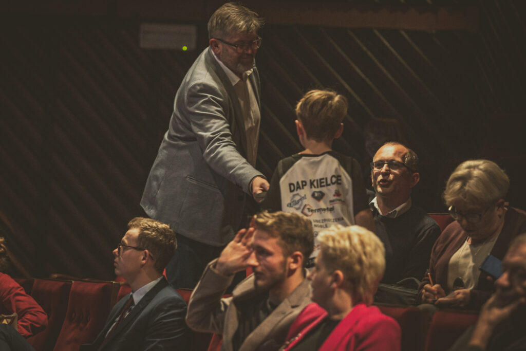 Zdjęcie. Mężczyzna w średnim wieku wyciąga melonik w stronę małego chłopca, stojącego między widxami. Chłopiec ma na koszulce z tyłu napis DAP Kielce.