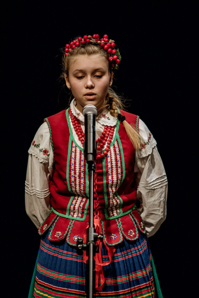 Zdjęcie. Blondynka w stroju ludowym w czerwonym serdaku śpiewa do mikrofonu.