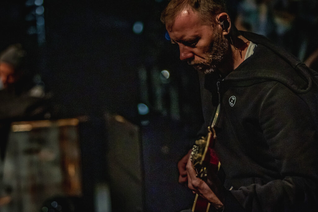 Mężćzyzna z brodą w czarnej bluzie z kapturem gra na gitarze. To muzyk z zespołu LemON.