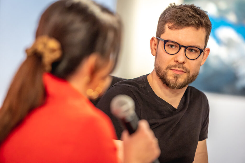 Zdjęcie. Dwie osoby rozmawiają ze sobą. Przodem do obiektywu młody mężczyzna w okularach i z brodą. To Maciej Makselon. Tyłem - kobieta w czerwonej marynarce z ługimi ciemnymi włosami. To Magdalena Fudala. Zdjęcie z "Salonu kobiet" - spotkanie z Maciejem Makselonem.