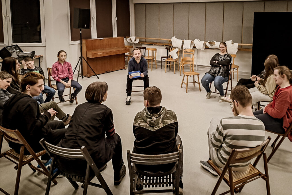 Zdjęcie. Uczestnicy warszratów teatralnych siedzą w półokręgu. Naprzeciwko nich aktor Wojciech Niemczyk.