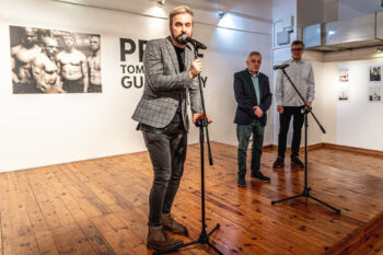 Zdjęcie. Wernisaż wystawy Proof Tomasza Gudzowatego. Na pierwszym planie wicedyrektor Artur Wijata mówi do mikrofonu. W tle fotografia Gudzowatego i dwóch mężczyzn.