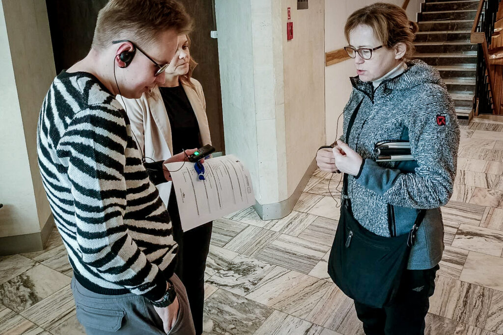 Zdjęcie. Kobieta i mężczyzna stoją przed wejściem na Małą Scenę KCK. Kobieta ma pod pachą złożóną białą laskę. Mężczyzna ma na uchu słuchawkę od sprzętu do audiodeskrypcji.