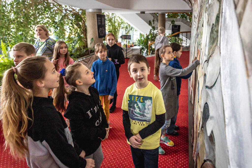 Zdjęcie. Grupa dzieci we Foyer Dużej SCeny KCK. Dzieci przyglądają się płaskorzeźbie przedstawiającej fantastyczne drzewo.