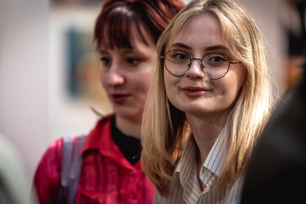 Zdjcie. Dwie młode dziewczyny podczas wernisażu "Dyplom". Blondynka w okularach uśmiecha się do obiektywu.