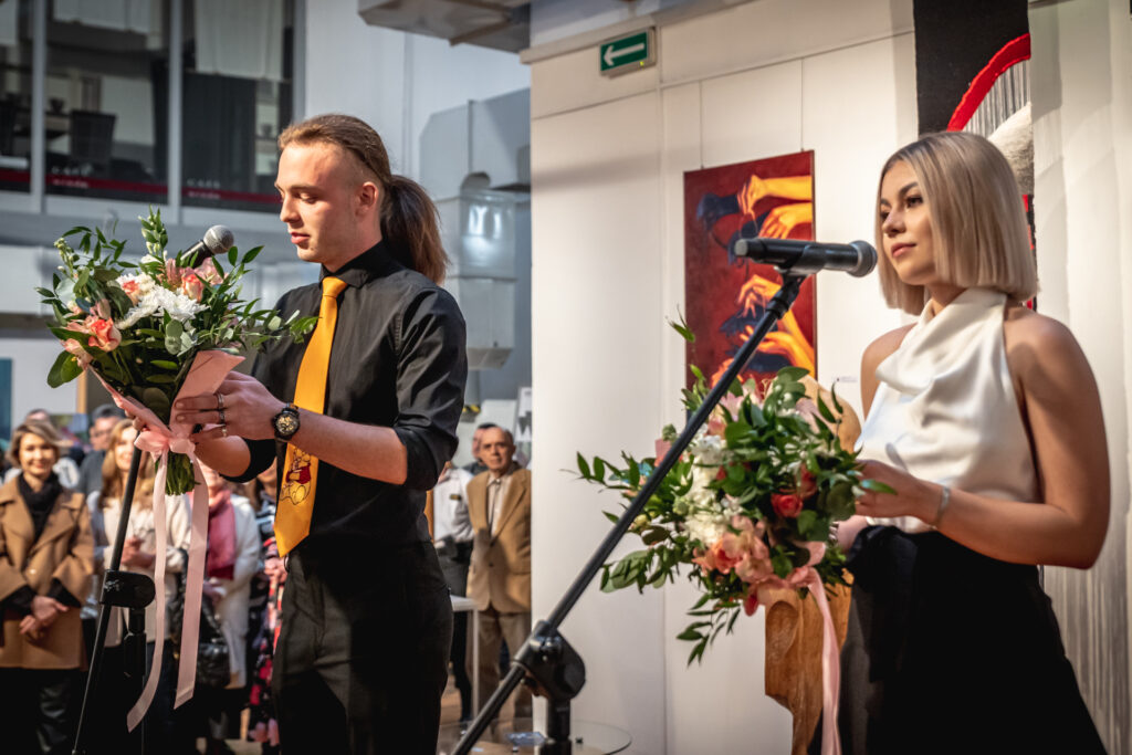 Zdjęcie. Dwójka młodych ludzi - chłopak i dziewczyna mówią do mikrofonów. Oboje trzymają w dłoniach bukiety kwiatów.