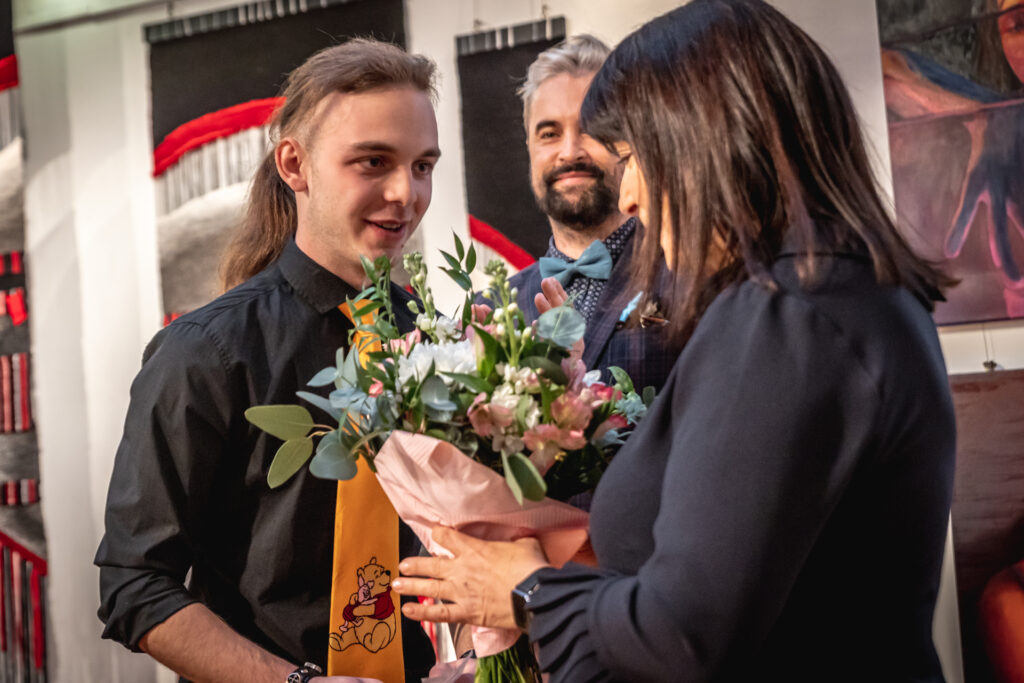 Zdjęcie. Wernisaż wystawy Dyplom. Młody mężczyzna wręcza bukiet kwiatów kobiecie o ziemnych prostych włosach. W tle wicedyrektor Artur Wijata.