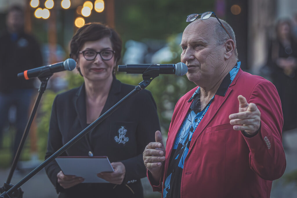 Zdjęcie. Dwie osoby stoją przy mikrofonach. Mężczyzna w czerwonej marynarce i niebieskiej koszuli w czarne kwiatowe desenie mówi do mikrofonu. To jazzman Włodek Pawlik. Z tyłu stoi ubrana w czarną marynarkę swzczupła kobieta o krótkich siemnych wlosach. Na nosie maokulary w ciemnych oprawkach. To dyrektor KCK Augustyna Nowacka.