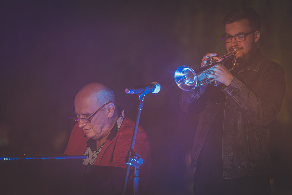 Zdjęcie. Spontaniczny koncert Włodka Pawlika podczas otwarcia napisu. Pawlki gra na pianinie, nad nim stoi Kuba Kowalski grający na trąbce.