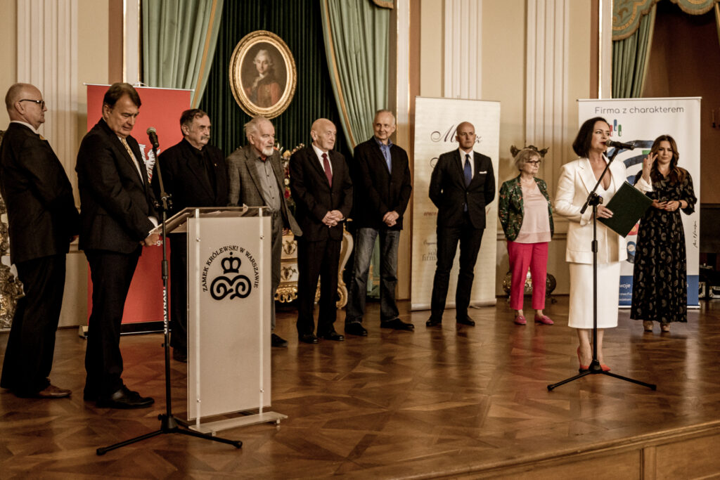 Zdjęcie. Dojrzali ludzie stoją w pólokręgu na scenie. Przy mikrofonie kobieta w białym kostiumie. To Jolanta Fajkowska.