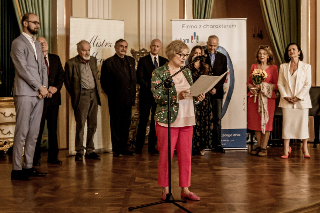 Zdjęcie. Kobieta o jasnych wlosach w okularach czyta nominację na scenie. W tle grupa ludzi.