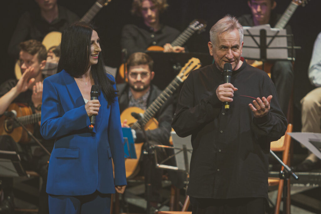 Zdjęcie. Jerzy Pikor mówi do mikrofonu. Obok niego kobieta o dlugich prostych ciemnych włosach w niebieskim kostiumie trzyma mikrofon. Patrzy w kierunku Jerzego Pikora. W tle - gitarowa orkiestra.