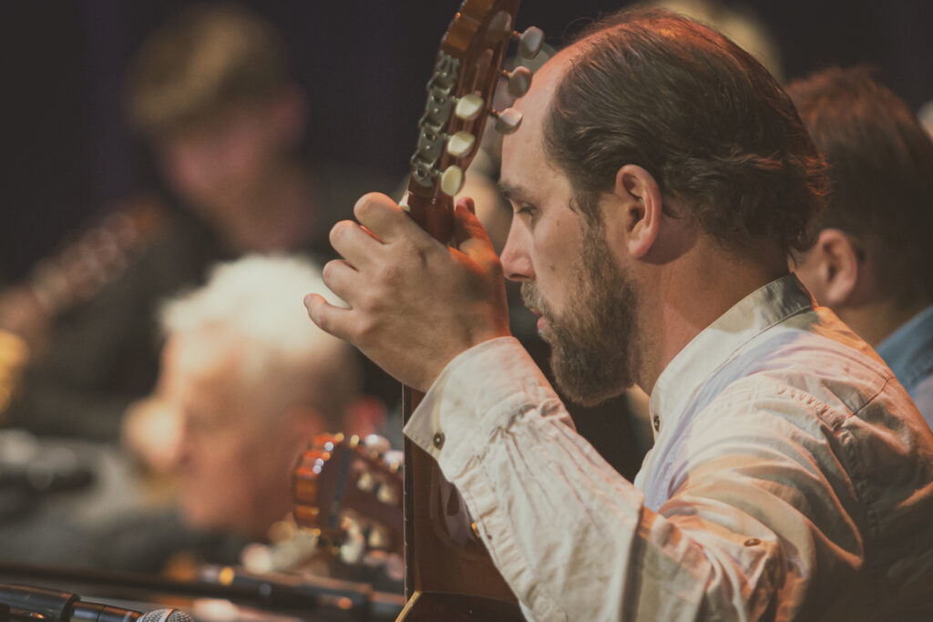 Zdjęcie. Łysawy gitarzysta z brodą gra na instrumencie.