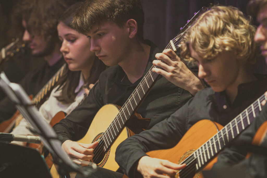 Zdjęcie. Czterech mlodych gitarzystów: trzech chłopaków i jedna dziewczyna gra na gitarach.