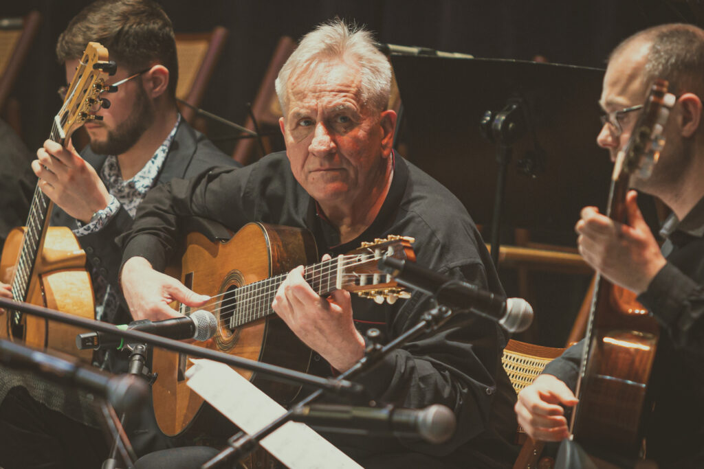 Zdjęcie. Jerzy Pikor gra na gitarze. Obok inni gitarzyści.