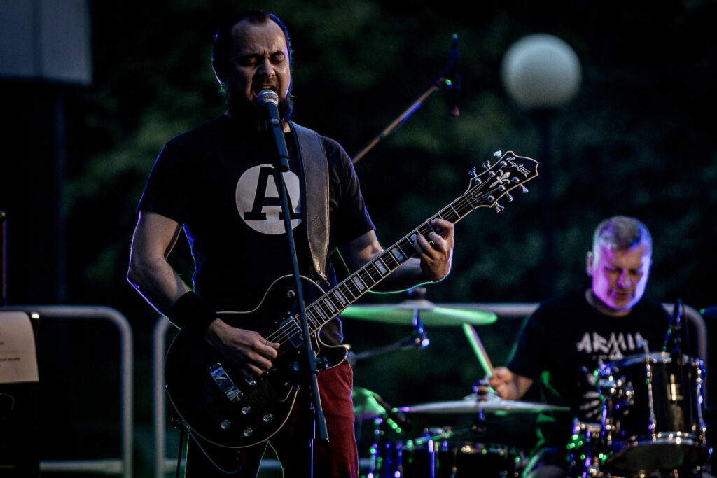 Zdjęcie. Brodaty gitarzysta gra i śpiewa do mikrofonu na letniej scenie KCK.