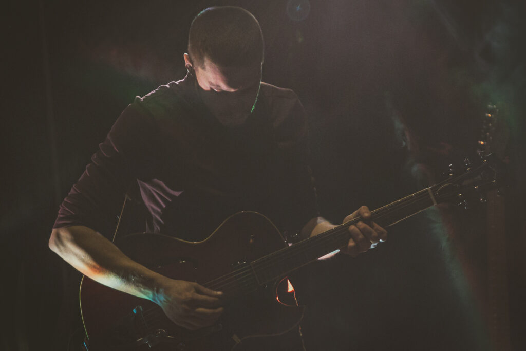 Zdjęcie. Krótko obcięty mężczyzna we fryzurze na jerzyka gra na gitarze. Scenę spowija ciemność, widać tylko kilka delikatnych refleksów świetlnych.