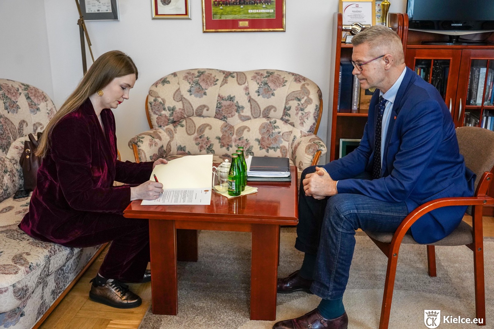 Zdjęcie. Młoda kobieta i wysoki mężczyzna siedzą po dwóch stronach stolika kawowego. Kobieta podpisuje dokument. Mężczyzna przygląda się jej. To dyrektor KCK Agata Klimczak-Kołakowska i prezydent Kielc Bogdan Wenta.