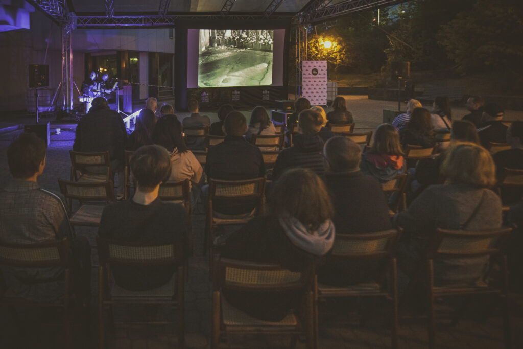 Zdjęcie. Widzowie oglądają czarno-biały film na dużym ekranie pod dachem sceny plenerowej KCK. Po lewej stronie od ekranu stoją dwaj muzycy.