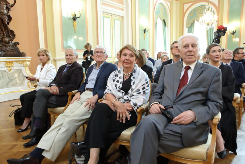 Zdjęcie. Rzędy eleganckich, historycznych krzeseł, na których siedzą widzowie. W pierwszym rzędzie siedzi trzech panów i trzy panie. Panowie są starsi i mają na sobie garnitury. Panie to dwie blondynki, jedna w czarnej suki z jasną chustą zawiązana na ramionach, druga ma na sobie biała bluzkę. W dłoniach trzyma telefon komórkowy.